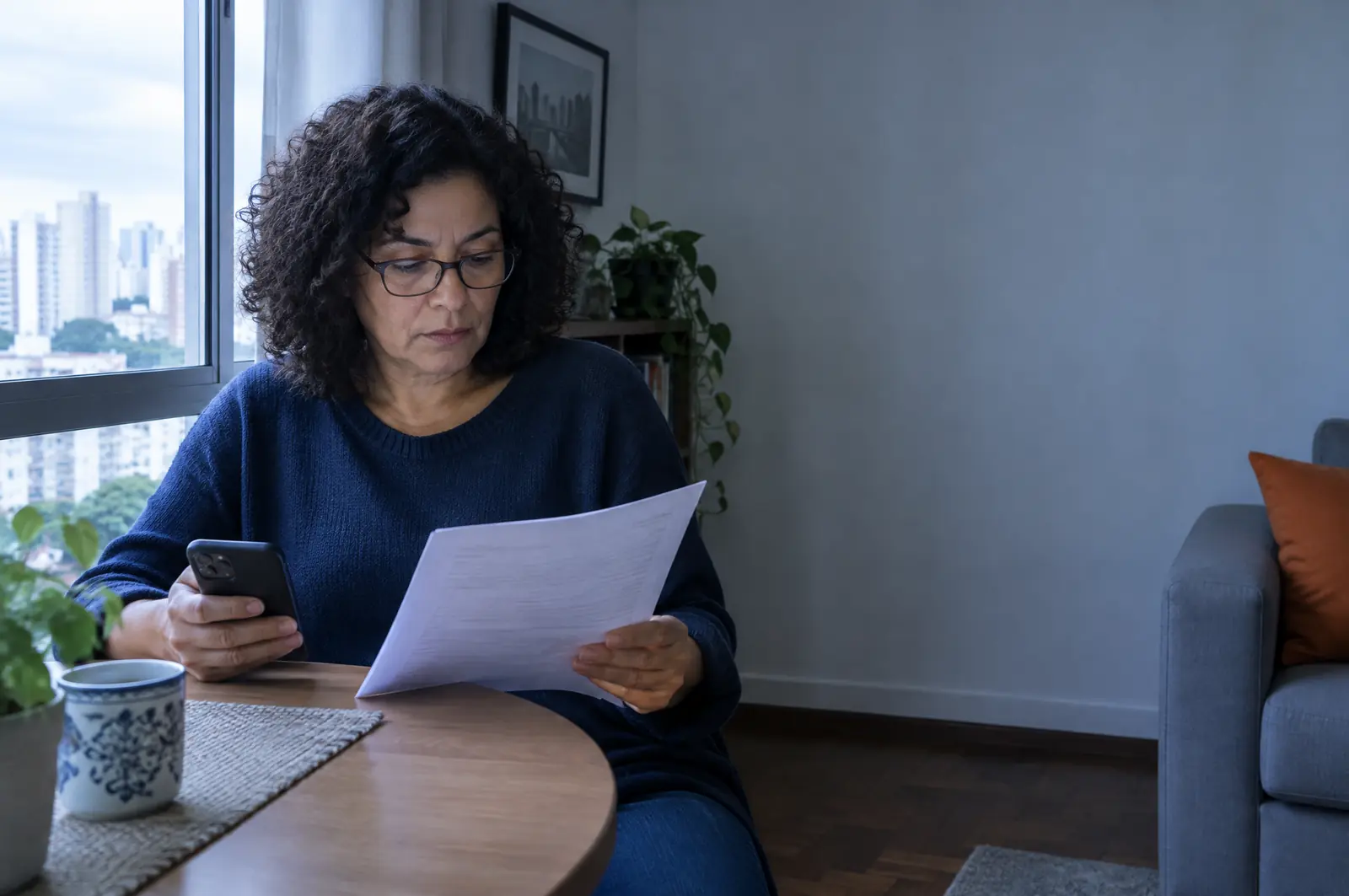 Mulher lê com atenção um documento impresso na sala de casa, com o celular na outra mão.